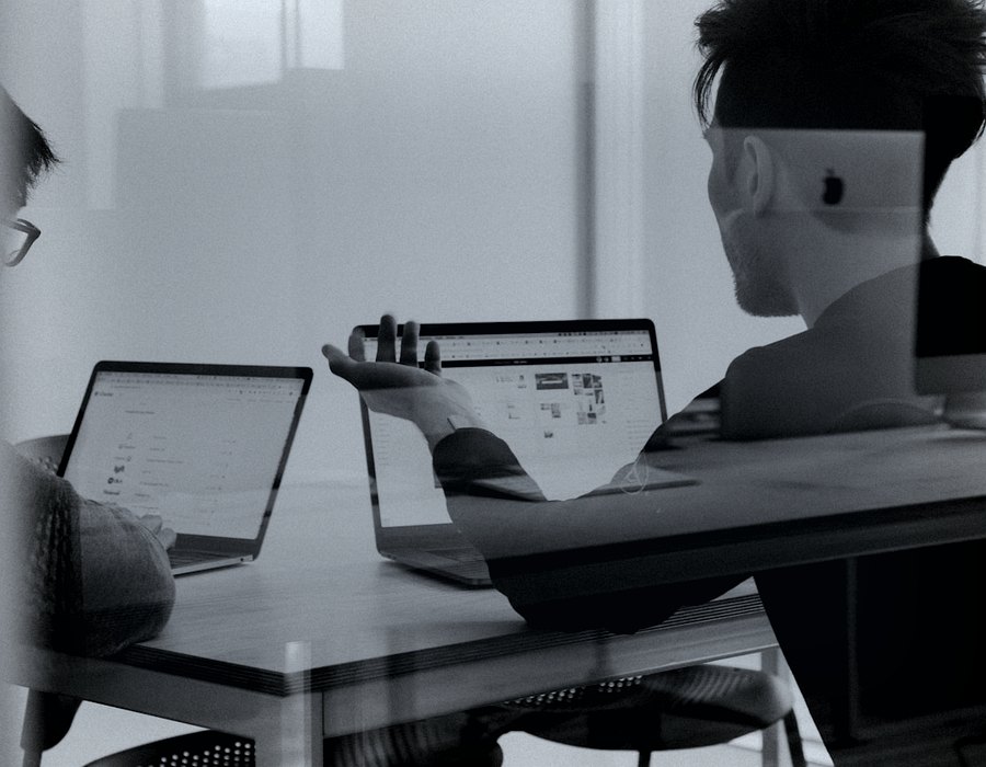 Two people reviewing work on laptops, seen through a glass partition